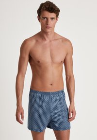 Blauwe zwembroek met een geometrisch patroon. Gemaakt van stof met een elastiche tailleband. Midden boven de knie lengte met een ontspannen pasvorm.