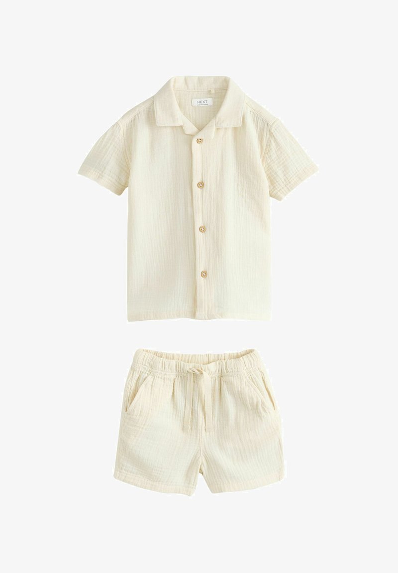 Camisa de manga corta color crema con botones, combinada con pantalones cortos con cintura elástica y cordón a juego, ambos hechos de tela ligera y texturizada.