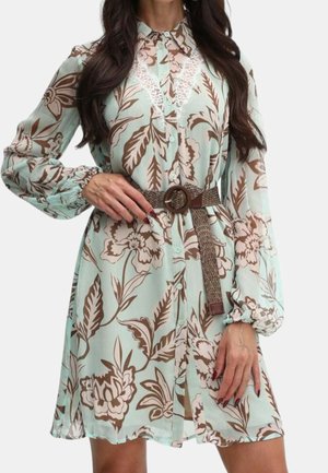 Frau trägt ein mintgrünes Blumenkleid mit Spitzen-Details, langen Puffärmeln, Knopfleiste vorne und einem braunen gewebten Gürtel mit runder Schnalle.