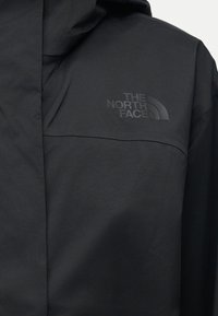 Posnetek črne nepremočljive jakne z kapuco in teksturiranim logotipom "The North Face" na prsih.