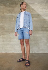 Hele sinine denim jope, valge T-särk, kulumisega denim lühikesed püksid ja mustad rihmadega sandaalid. Tellismüür ja betoonpõranda taust.