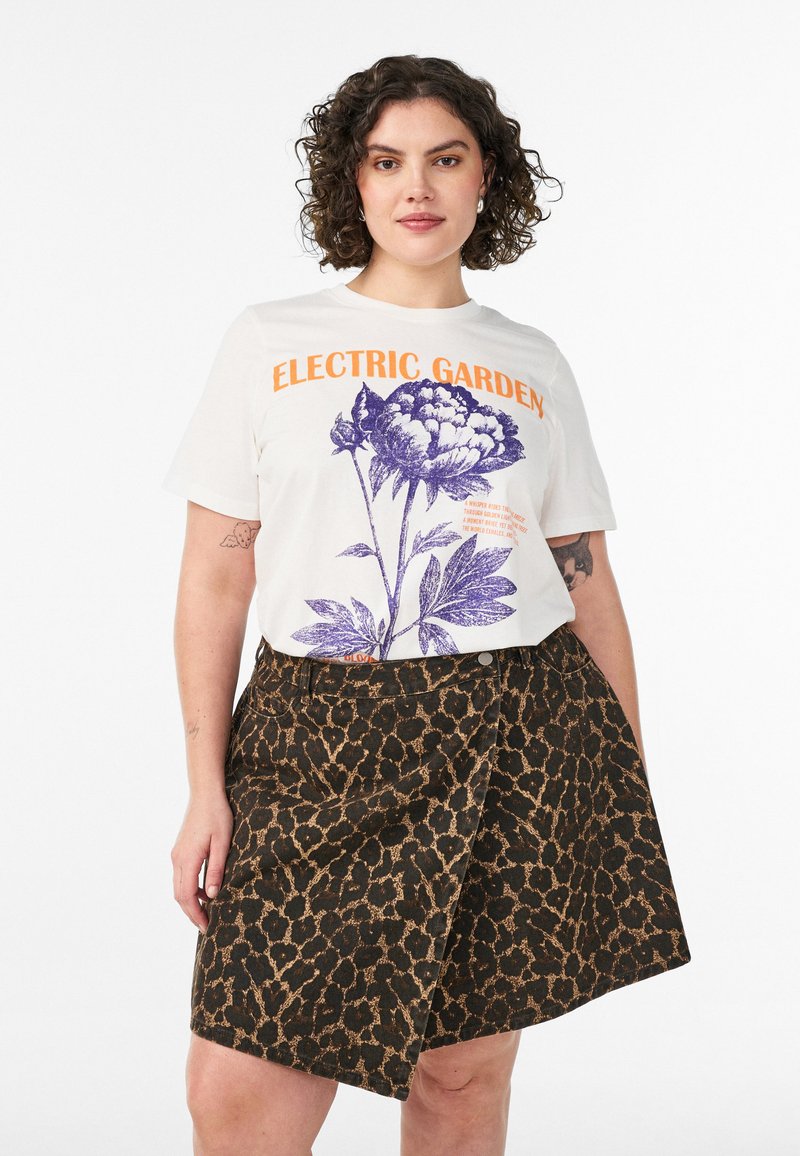 Γυναίκα με σγουρά μαλλιά που φοράει λευκό T-shirt "Electric Garden" με μωβ floral σχέδιο και καφέ φούστα με λεοπάρ σχέδιο.