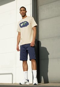 T-shirt bege com uma graphic azul e verde, combinada com calções azul marinho. Meias desportivas brancas e sapatilhas brancas completam o conjunto.