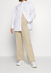 Chemise blanche boutonnée à la coupe décontractée et manches longues, associée à un pantalon large beige à fines rayures et des baskets blanches.