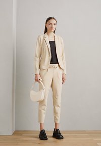 Blazer beige ajusté et pantalon assorti sur un haut noir ; femme tenant un sac à main crème arrondi ; mocassins noirs ; texture lisse.