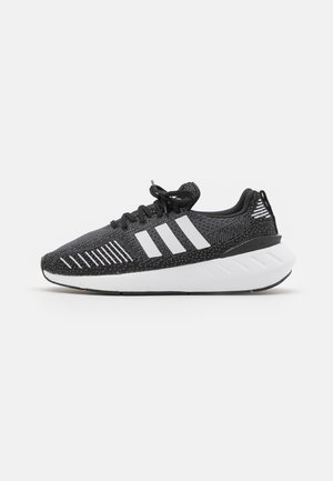 Chaussures de sport noires avec un dessus texturé, design à trois bandes blanches, semelle en caoutchouc et accents blancs contrastants sur le talon.