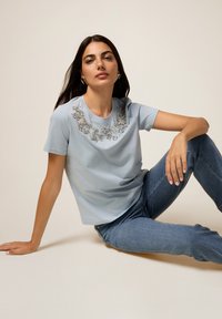 Chemise bleu clair à manches courtes avec des embellissements en perles florales au niveau de l'encolure, associée à un jean bleu à jambe droite. Texture lisse.