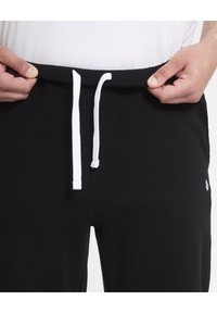 Pantalon de jogging noir avec une large ceinture élastique, cordon de serrage blanc et petit logo près de l'ourlet. Matière en coton, texture douce.