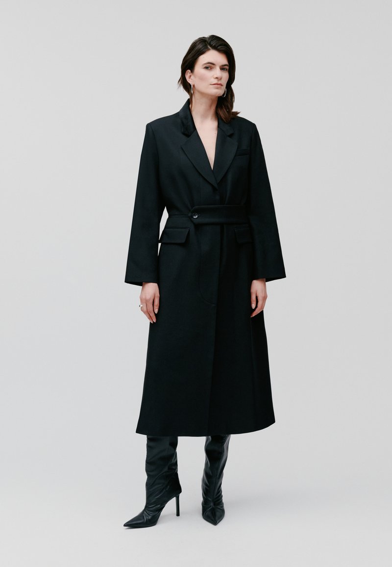 Cappotto lungo nero con un design strutturato, ampie revers, vita con cinturino e due tasche frontali. Abbinato a stivali neri a punta fino al ginocchio.