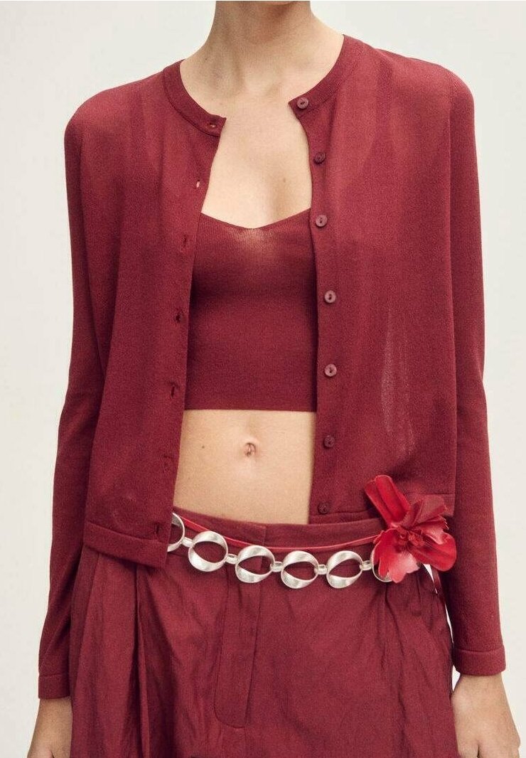 Femme portant un cardigan bordeaux sur un crop top assorti, un pantalon taille haute et une ceinture à chaîne argentée avec un accessoire fleur rouge.