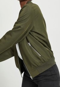 Veste bomber vert olive en tissu doux, avec une poche zippée sur le côté gauche et des poignets et un ourlet côtelés.