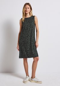 Bezramionowa zielona sukienka w czarny wzór lamparta, luźny krój, do kolan, zestawiona z białymi i beżowymi sneakersami.