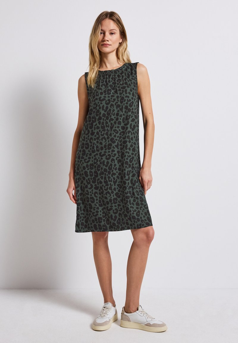 Bezramionowa zielona sukienka w czarny wzór lamparta, luźny krój, do kolan, zestawiona z białymi i beżowymi sneakersami.