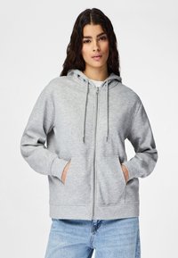Mujer joven con sudadera con capucha gris claro con cremallera y bolsillos, y vaqueros azul claro, de pie frente a un fondo blanco liso.