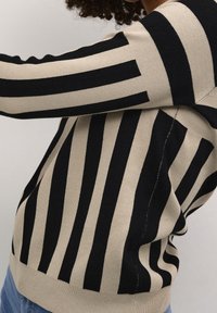 Person iført en beige og sort stribet sweater med skiftende lodrette og vandrette striber, kombineret med blå jeans.