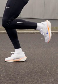 Nike running shoes in white with blue and orange accents. Lightweight material, cushioned sole, and a sleek design. Black athletic leggings visible.