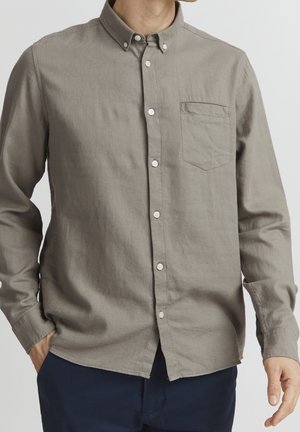 Hombre con una camisa marrón clara de manga larga con botones y bolsillo en el pecho, y pantalones azul oscuro, con las manos parcialmente en los bolsillos.