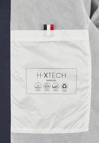 Witte regenjas met mesh details, voorzien van een label met iconen voor waterdicht, ademend, winddicht en gerecycled materiaal.