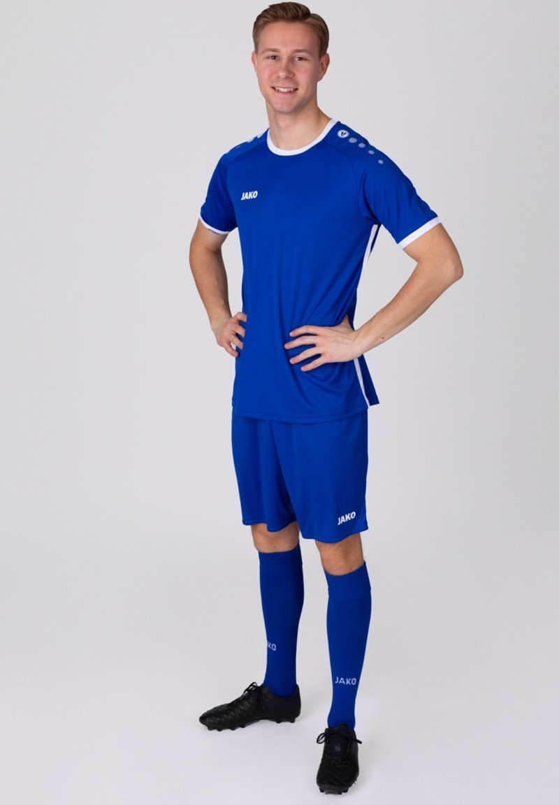 Completo da calcio blu realizzato in materiale leggero; presenta accenti bianchi, logo sul petto e calzettoni abbinati. Il modello sta in piedi con sicurezza.