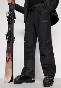 Fekete sínadrág különböző textúrákkal és nagy cargo zsebekkel, fekete sícipőkkel párosítva. Egy snowboard kötéssel a közelben van tartva.