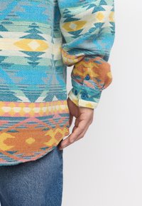 Kleurrijk geweven shirt met blauwe, gele en oranje geometrische patronen, lange mouwen, gestructureerde stof en blauwe knopen op de manchetten.
