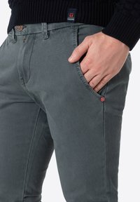 Graue, strukturierte Hose mit schmaler Passform, ausgestattet mit einer Seitentasche und einem kleinen roten Logo-Detail am Bund. Gestickte Akzente in der Nähe der Taschen.