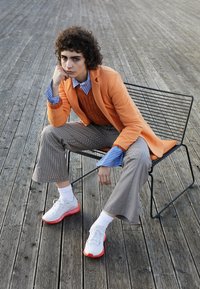 Blazer orange sur un pull orange texturé, poignets de chemise rayés bleus, pantalon à carreaux et baskets blanches avec des accents rouges.