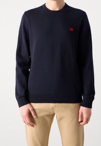 Granatowy sweter z dzianiny z okrągłym dekoltem, prążkowanymi mankietami i dołem, z małym czerwonym logo na klatce piersiowej. Gładka faktura.