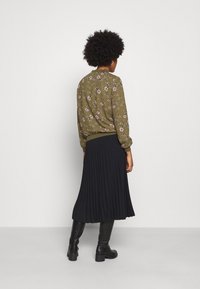 Olivenfarvet blomsterbluse med samlede manchetter, parret med en sort plisseret nederdel, båret med højhælede sorte støvler.