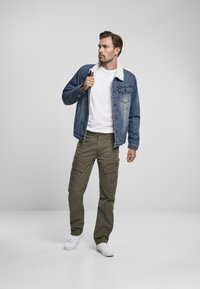 Giacca in denim con colletto in shearling, indossata su una maglietta bianca, abbinata a pantaloni cargo color oliva e sneaker bianche. Design semplice e casual.