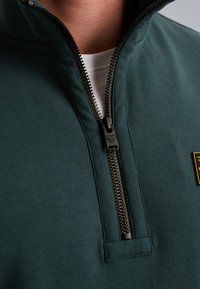 Dark green zip-up jacket with a short collar, featuring a black zipper and a golden logo patch on the chest. Smooth fabric texture.