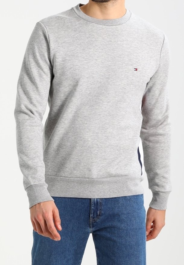 Homme portant un sweat-shirt gris clair à col ras du cou avec un petit logo Tommy Hilfiger sur la poitrine et un jean bleu.