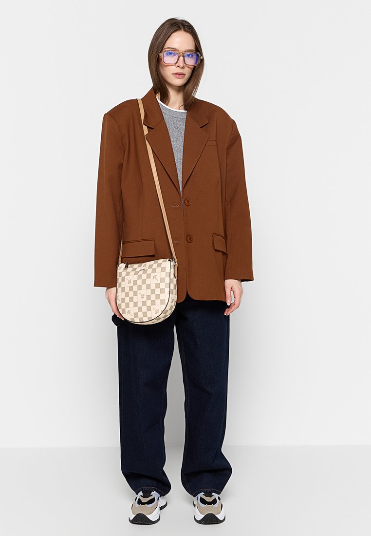 Übergroße braune Blazerjacke, grauer Pullover, dunkle weit geschnittene Jeans und weiße Sneaker. Cremefarbene Umhängetasche mit karierter Musterung. Blaue Brille.