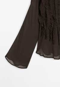 Blusa lunga a maniche trasparenti marrone con dettagli in raso verticali testurizzati sul fronte e maniche a campana su uno sfondo bianco.