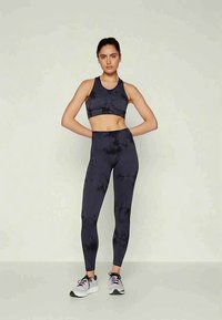 Mujer de pie con las manos detrás de la espalda, vestida con un sujetador deportivo negro de estilo tie-dye, leggings a juego y zapatillas blancas, frente a un fondo sencillo.