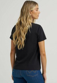 Camiseta negra de manga corta en algodón, con un ajuste clásico. Textura suave con una costura en el cuello y los hombros discreta; combinada con unos vaqueros azules.