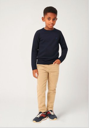 Marineblaues Strickpullover mit Rundhalsausschnitt, beigefarbene Jeanshose und marineblaue Sneakers mit roten und weißen Akzenten. Der Pullover hat ein kleines Logodetail.