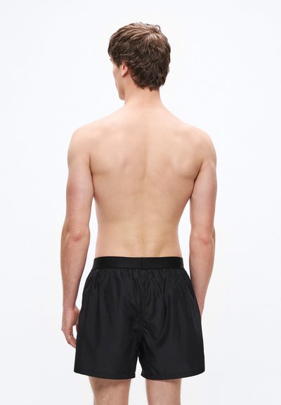 Pantalones cortos de baño negros con cintura elástica, tela ligera y ajuste relajado, mostrados desde una perspectiva trasera sobre un fondo neutro.