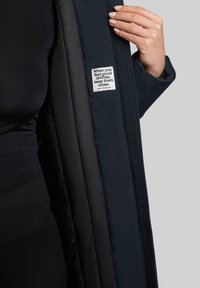 Mørkeblå dunjakke med en glat overflade, kviltet mønster og et mærke med tekst indeni. Lynlåsåbning og ribbede kanter er synlige.