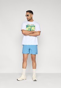 Λευκό γραφικό μπλουζάκι με πολύχρωμο σχέδιο φύσης, ανοιχτό μπλε σορτς, λευκές κάλτσες, και μπεζ αθλητικά παπούτσια. Σκούρα γυαλιά ηλίου συμπληρώνουν την εμφάνιση.