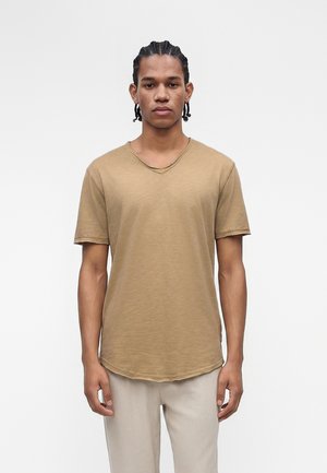 Jeune homme aux cheveux tressés debout, portant un t-shirt beige à manches courtes et un pantalon de couleur claire sur un fond blanc uni.