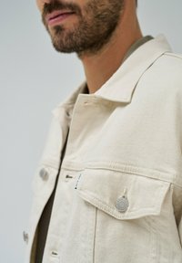 Chaqueta de mezclilla beige con cuello doblado, bolsillos frontales con detalles de botones plateados y una tela texturizada. Vista en primer plano sin fondo.