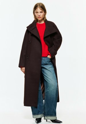 Jeune femme portant un long manteau sombre, un pull rouge, un jean bleu large et des chaussures noires pointues, debout avec une main dans sa poche.