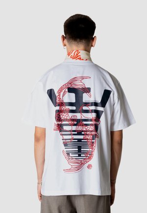 Mand med kort hår iført hvid oversized T-shirt med stort sort og rødt koi fisk grafik på ryggen, står mod en ensfarvet baggrund.