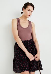 Top tank canelado castanho combinado com uma saia preta com pequenos padrões florais vermelhos. Modelo segura uma alça castanha, de pé contra um fundo neutro.