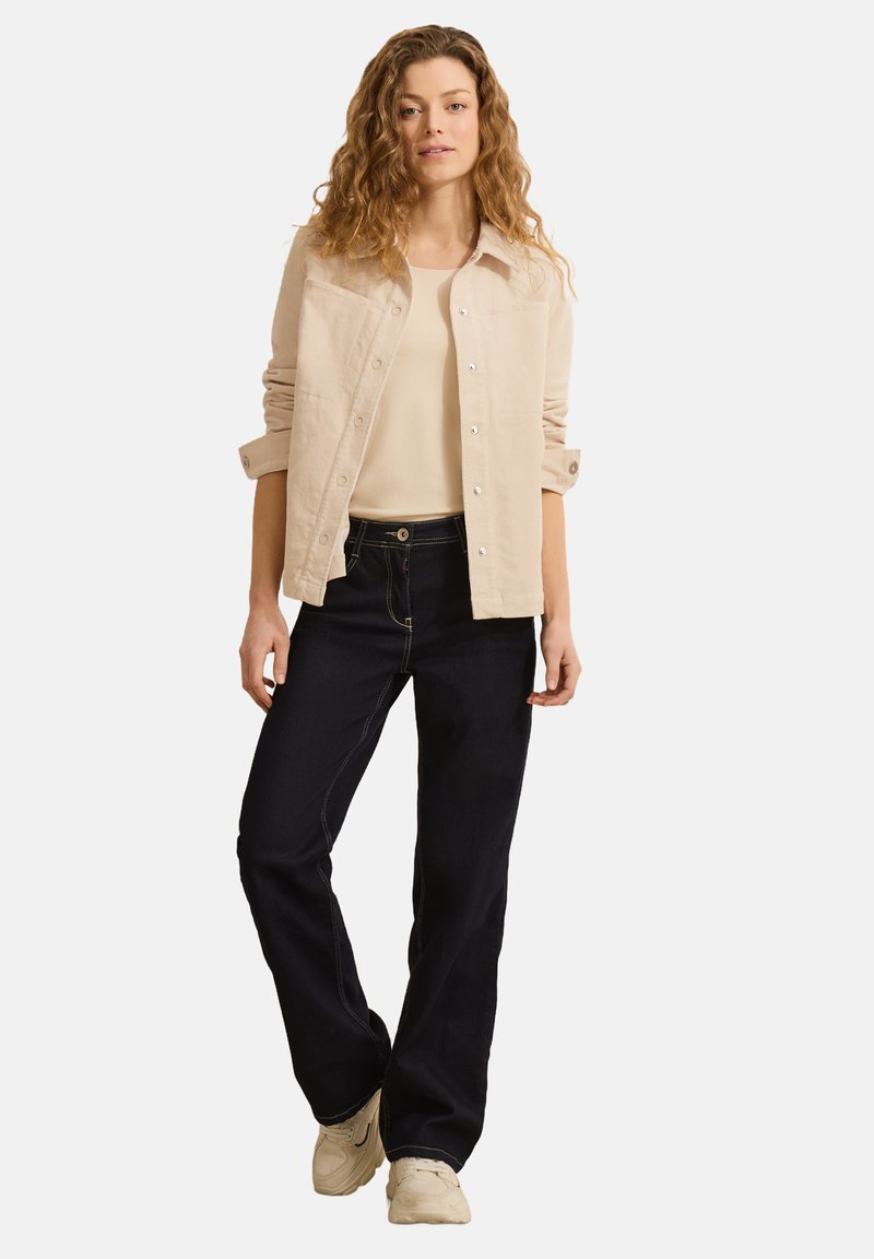 Beige knäppjacka, krämfärgad linne, mörka denimflared jeans, ljusa sneakers. Mjuk tyg, avslappnad passform, synliga sömmar på jeans.