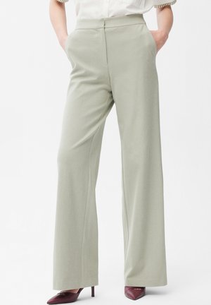 Donna con pantaloni a vita alta, gamba larga, di colore grigio chiaro con tasche frontali e tacchi a punta color bordeaux, mani nelle tasche.