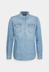 Ljusblå denimskjorta med långa ärmar, spetsig krage, två bröstfickor och metallknappar. Har stickad detaljer och en kurvad fåll.