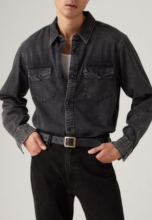 Hombre con una camisa de mezclilla negra desgastada sobre una camiseta blanca sin mangas, pantalones negros y cinturón negro con hebilla cuadrada dorada, con las manos en los bolsillos.