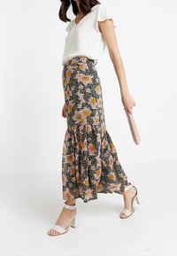 Frau, die in einer weißen Kurzarmbluse, einem schwarzen Maxirock mit orangefarbenen Blumen und beigen High Heels geht, während sie eine beigen Clutch hält.